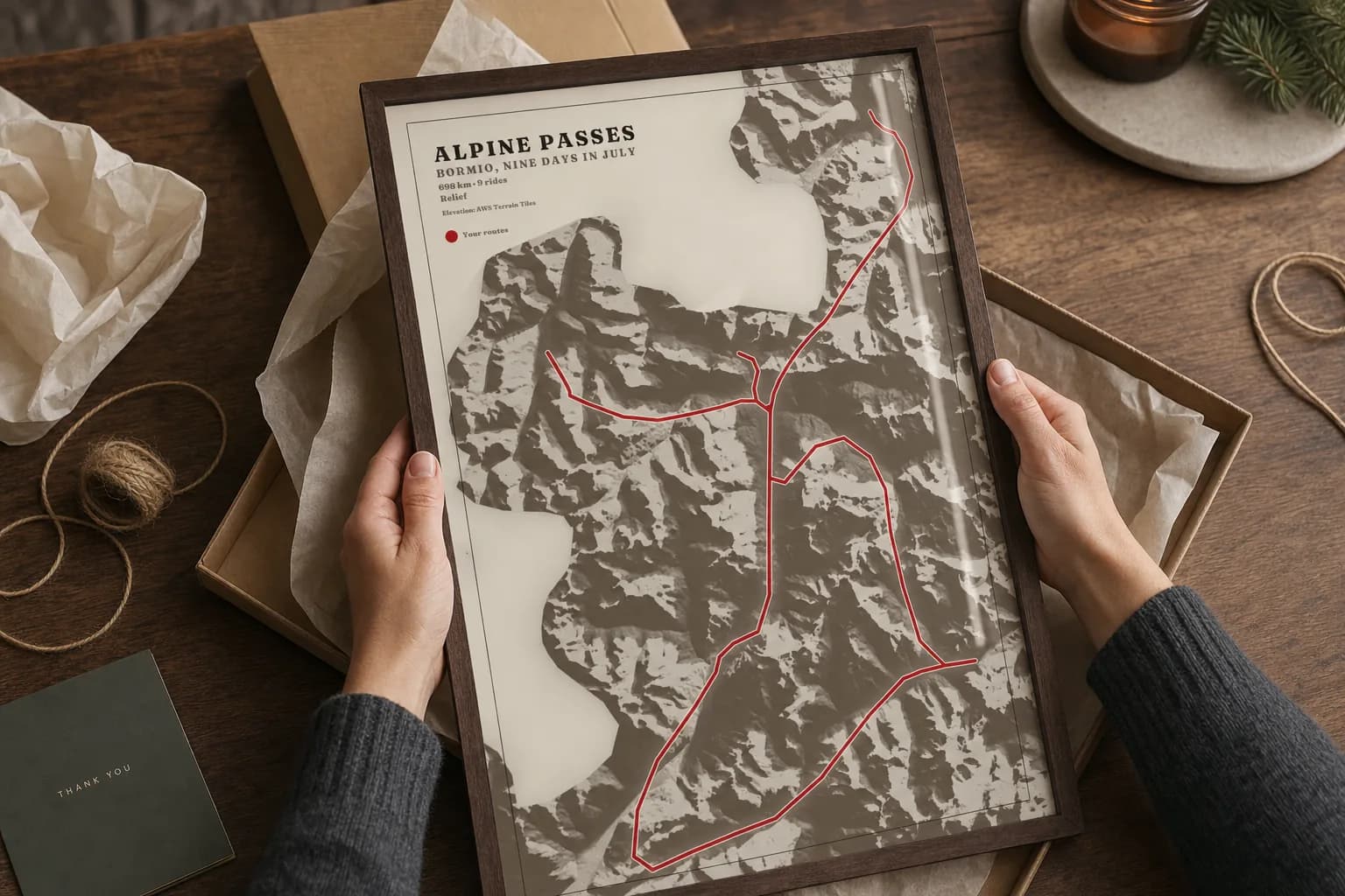 Hands lifting the framed Alpine passes poster out of an opened kraft gift box lined with tissue, twine and a card on the table beside it.