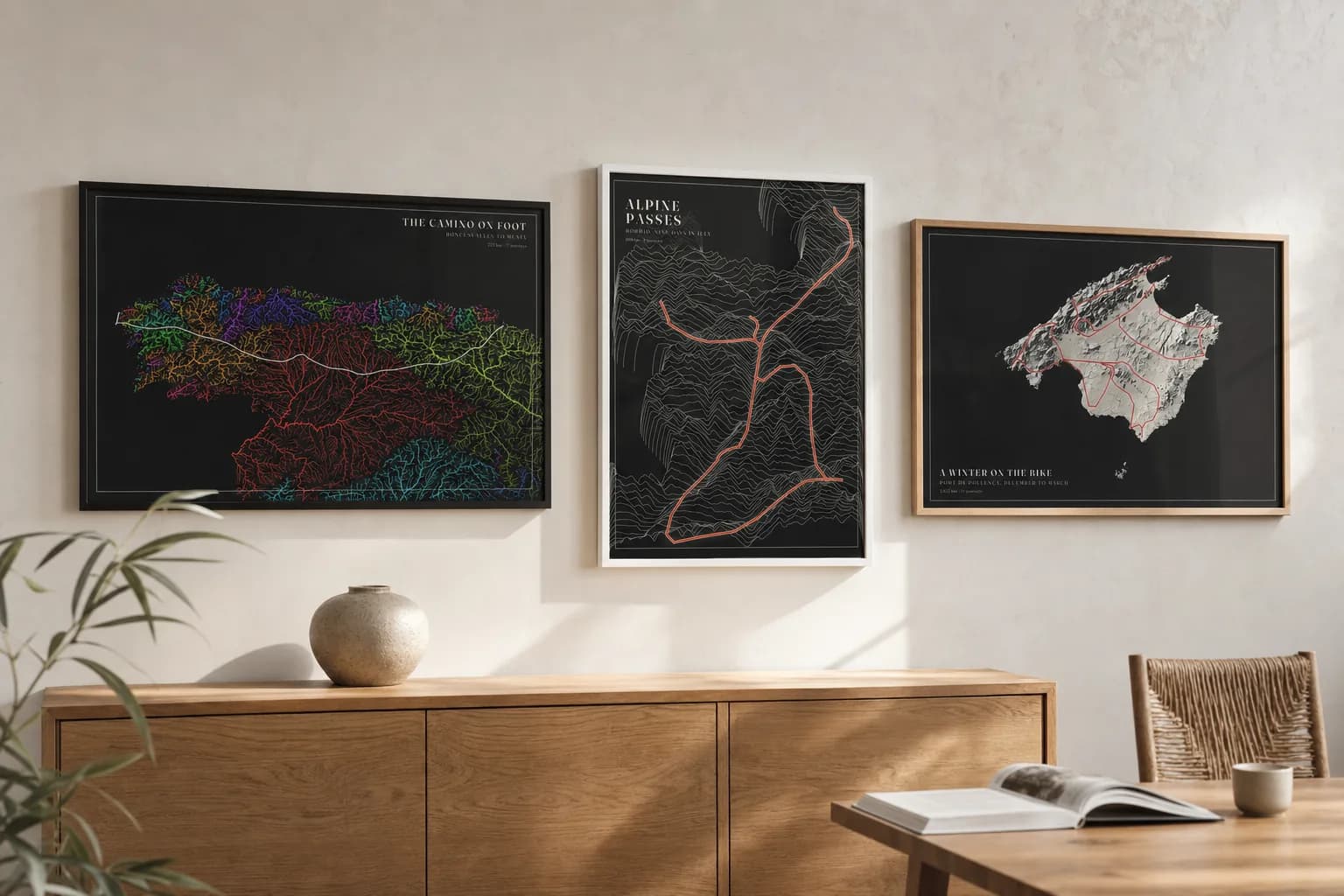 Three posters hung side by side above an oak sideboard: the Camino in bright River basins on black, the Alps in cream Ridgelines on black, and Mallorca in pale Plaster relief on black.